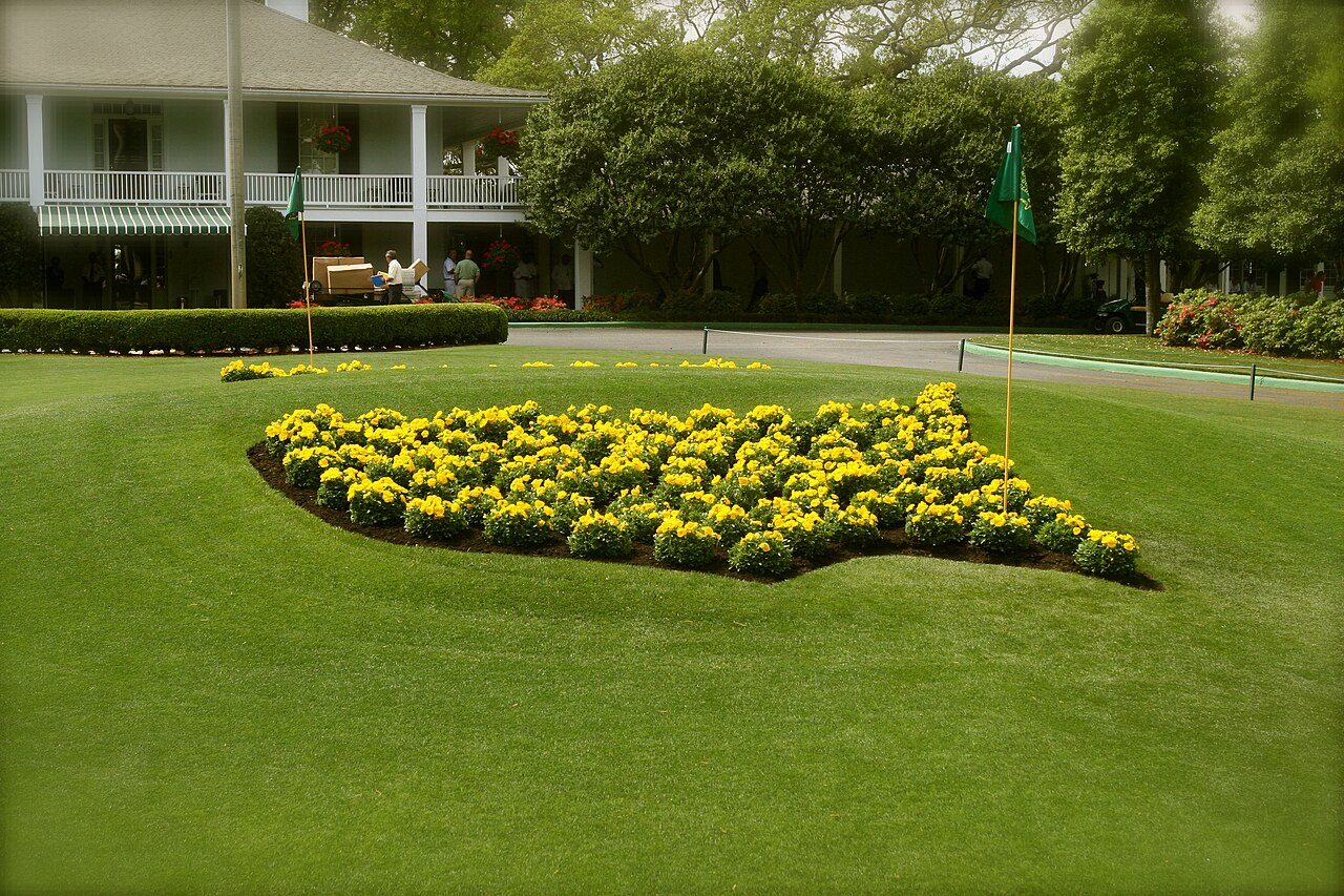 Yellow flowerbed planted in the shape of the Masters logo, with a flagstick, in front of the Augusta National clubhouse