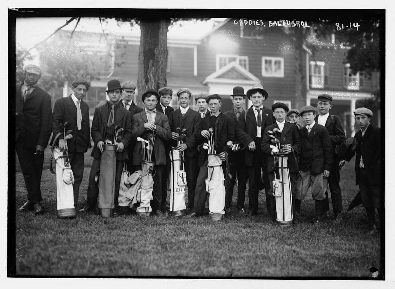 Black-and-white photograph of a row of caddies holding golf bags outside the clubhouse at Baltusrol