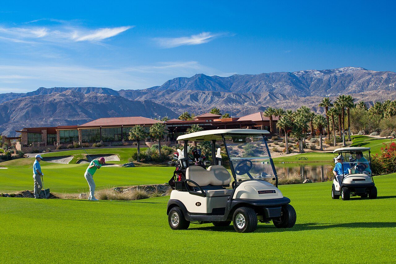 Two golf carts and a pair of golfers on a green desert resort course with mountains behind