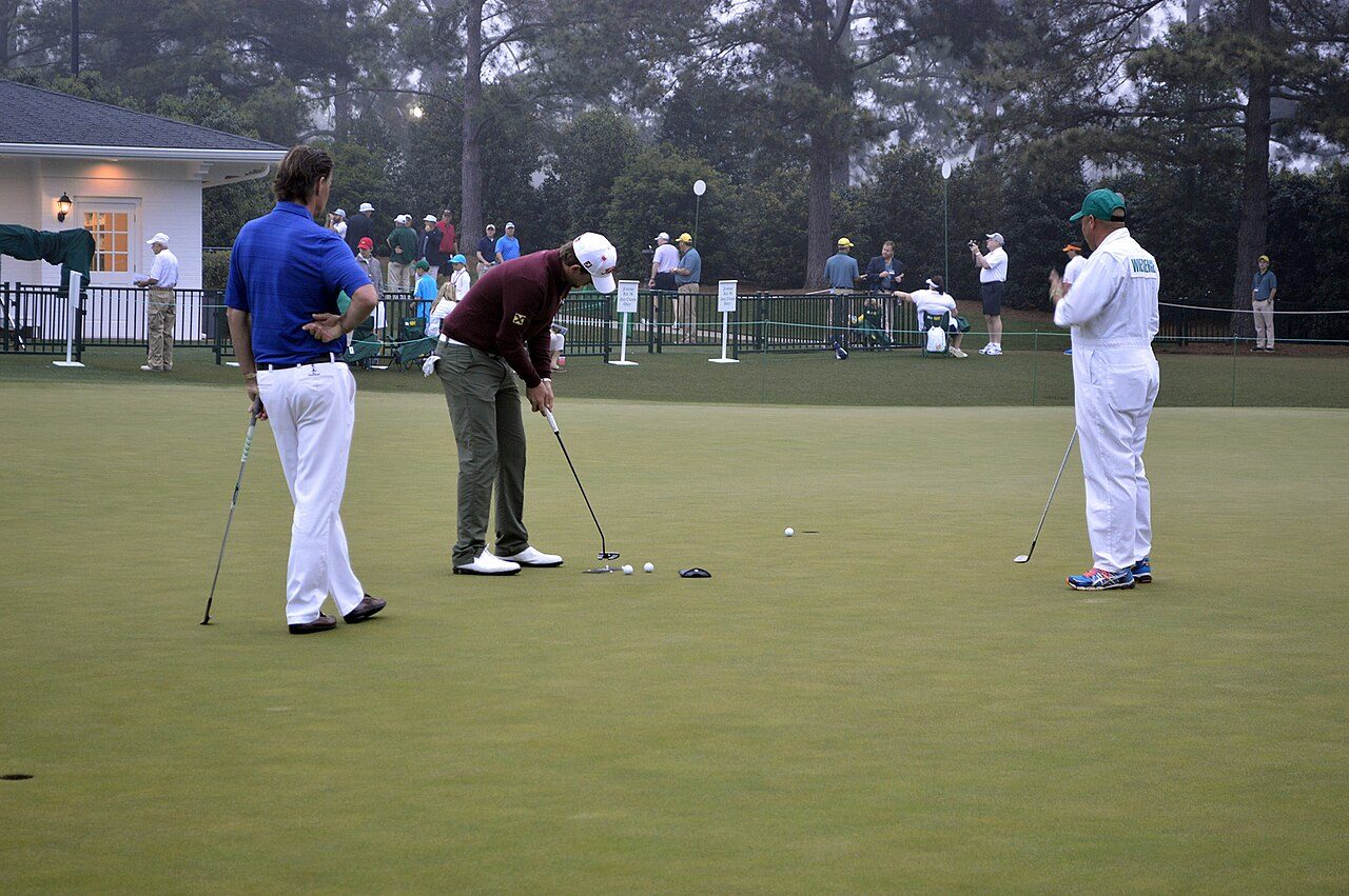 A player putting on the practice green at the 2015 Masters while his caddie in a white Augusta National jumpsuit looks on