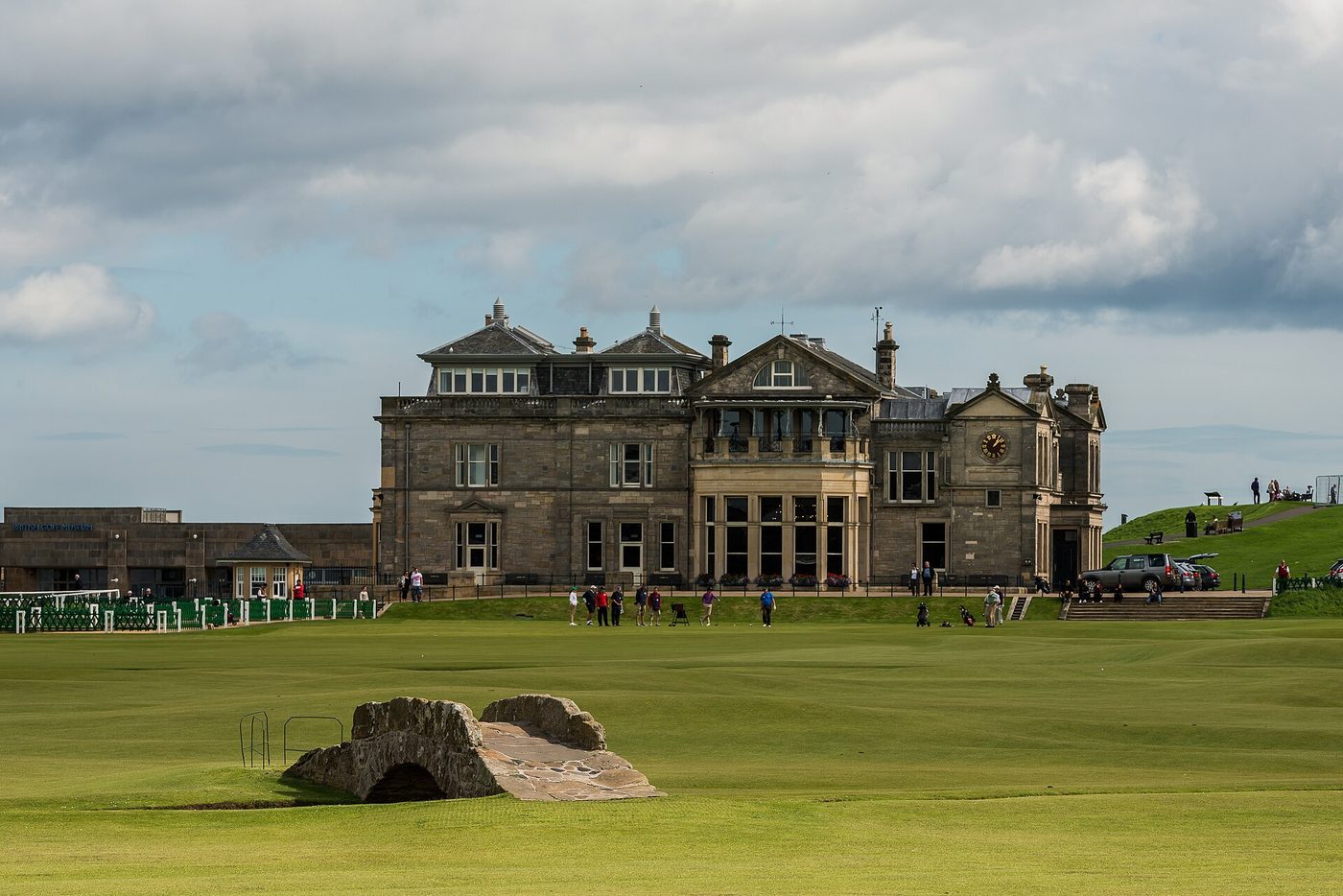 Golfers on the first tee of the Old Course at St Andrews in front of the R&A clubhouse, with the Swilcan Bridge in the foreground