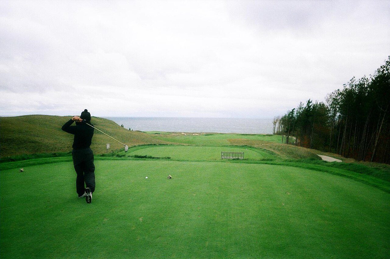 A golfer hitting a tee shot down a fescue-lined fairway toward Lake Michigan at Arcadia Bluffs