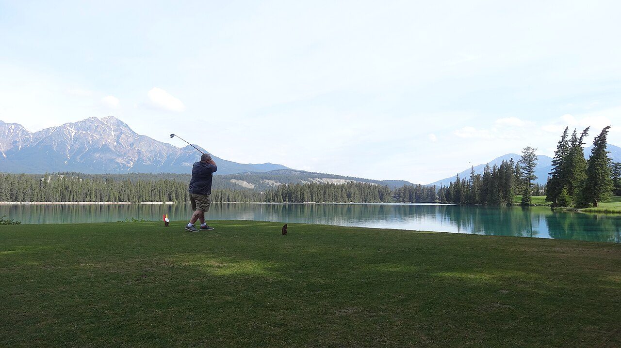 A golfer driving from a tee beside a lake below the mountains at Jasper Park Lodge in Alberta